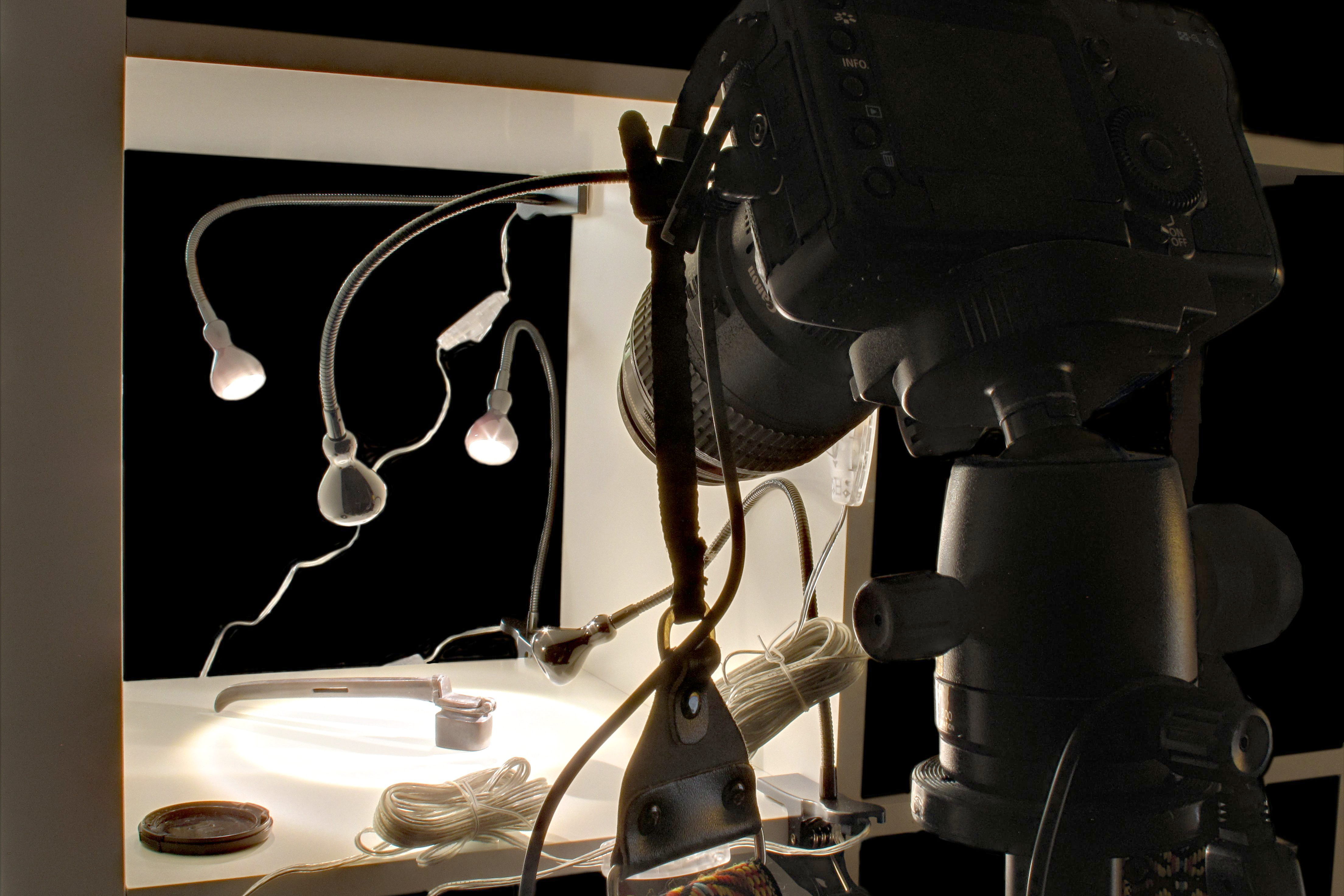 A camera on a tripod facing a lit shooting surface, with gooseneck lamps and coiled cables around it.