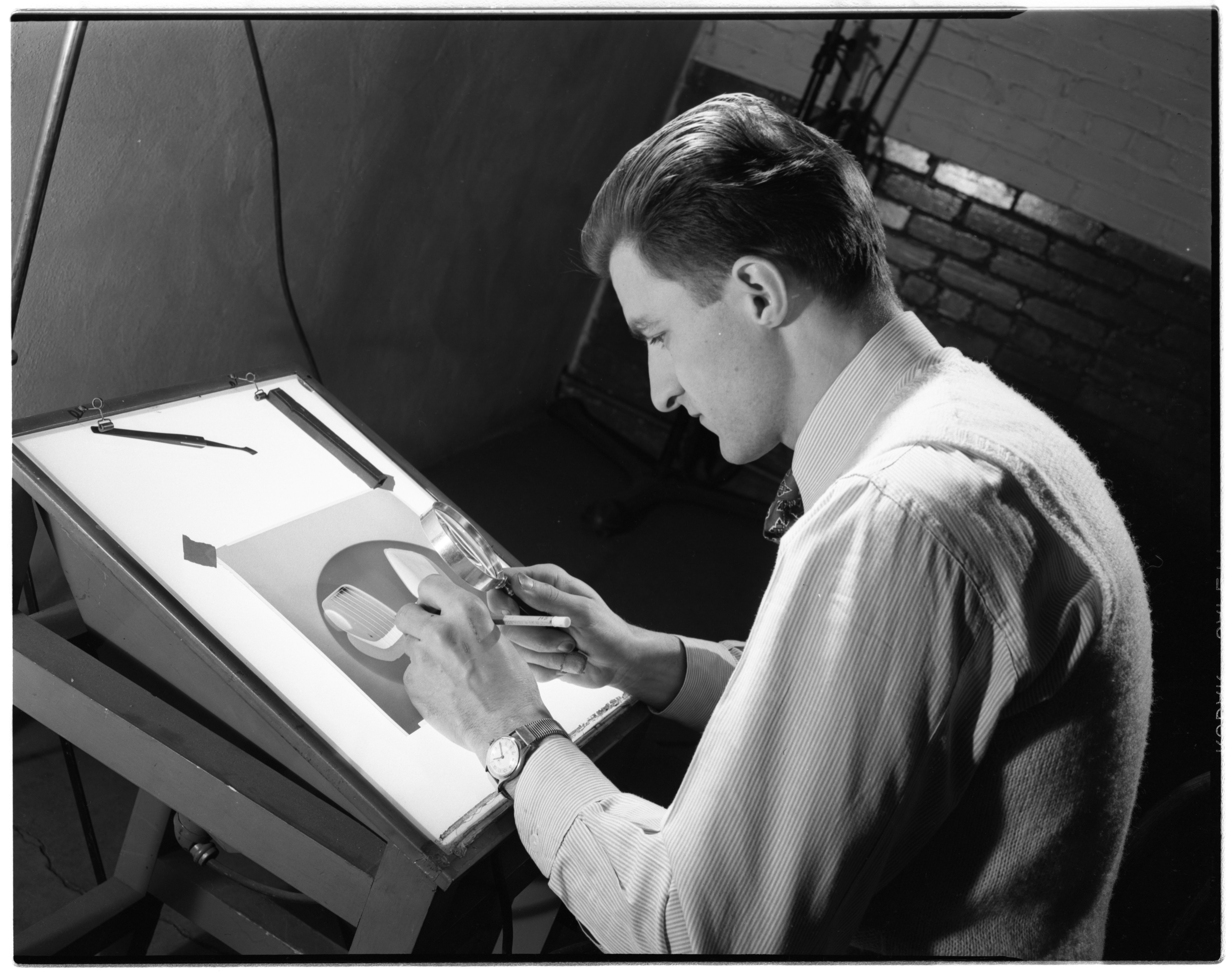 A man leaning over a backlit negative on a sloped desk, working with a loupe and a pencil.